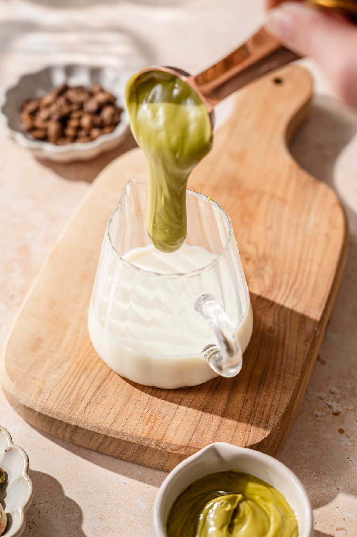 a hand pouring a tablespoon filled with pistachio cream into a pitcher of oat milk.