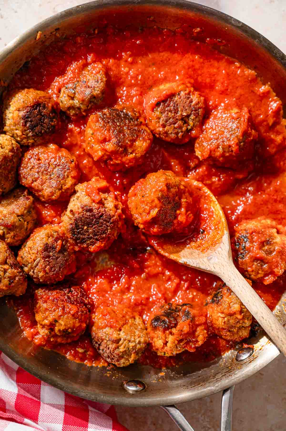 a wooden spoon scooping up a homemade vegan meatball in a stainless steel pan with marinara sauce.