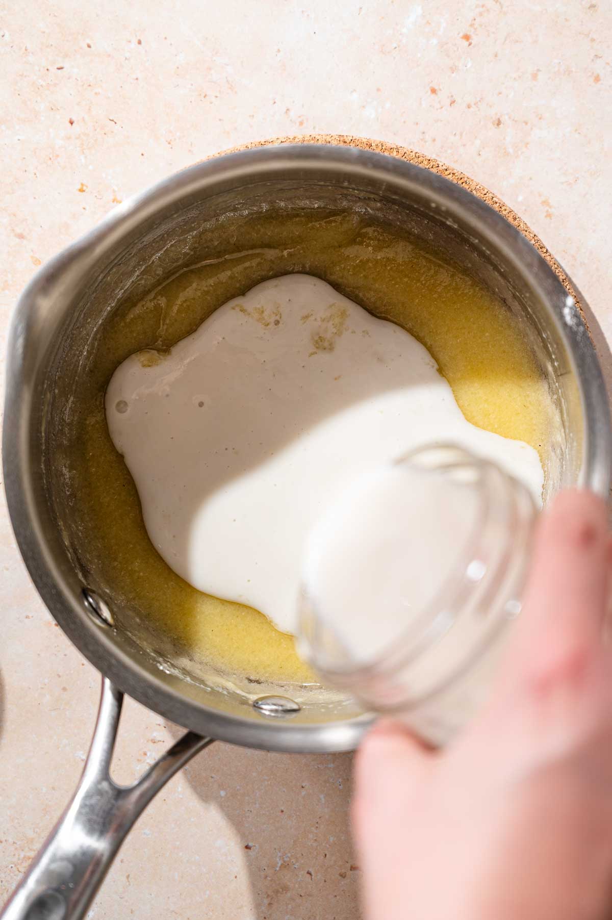 a hand pouring dairy free milk into the roux for the vegan cheese sauce.