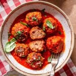 Vegan meatballs simmered in marinara sauce, garnished with fresh basil and parsley, served in a shallow bowl with crusty bread on the side.