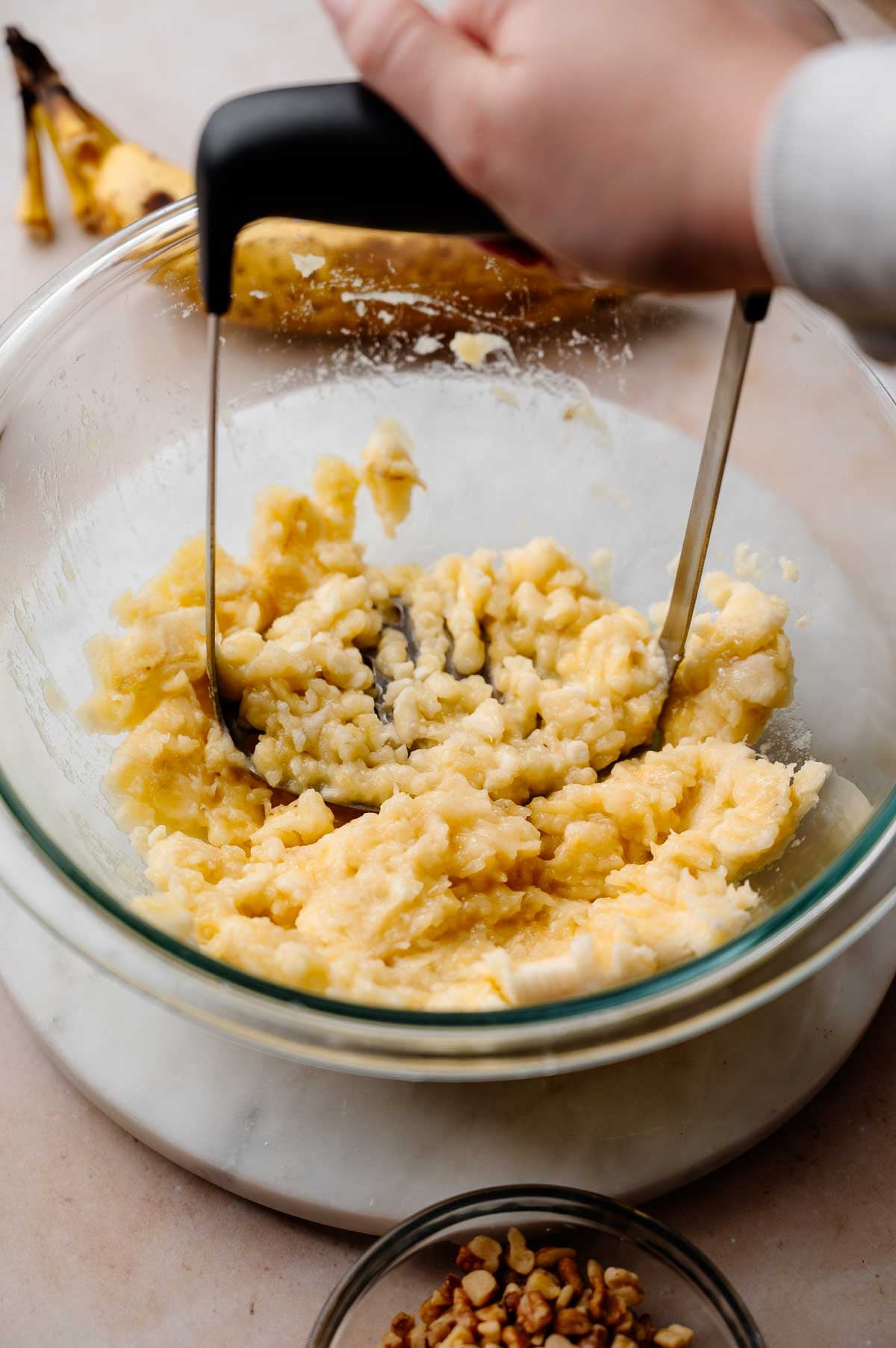 a hand mashing ripe bananas in a large glass bowl.