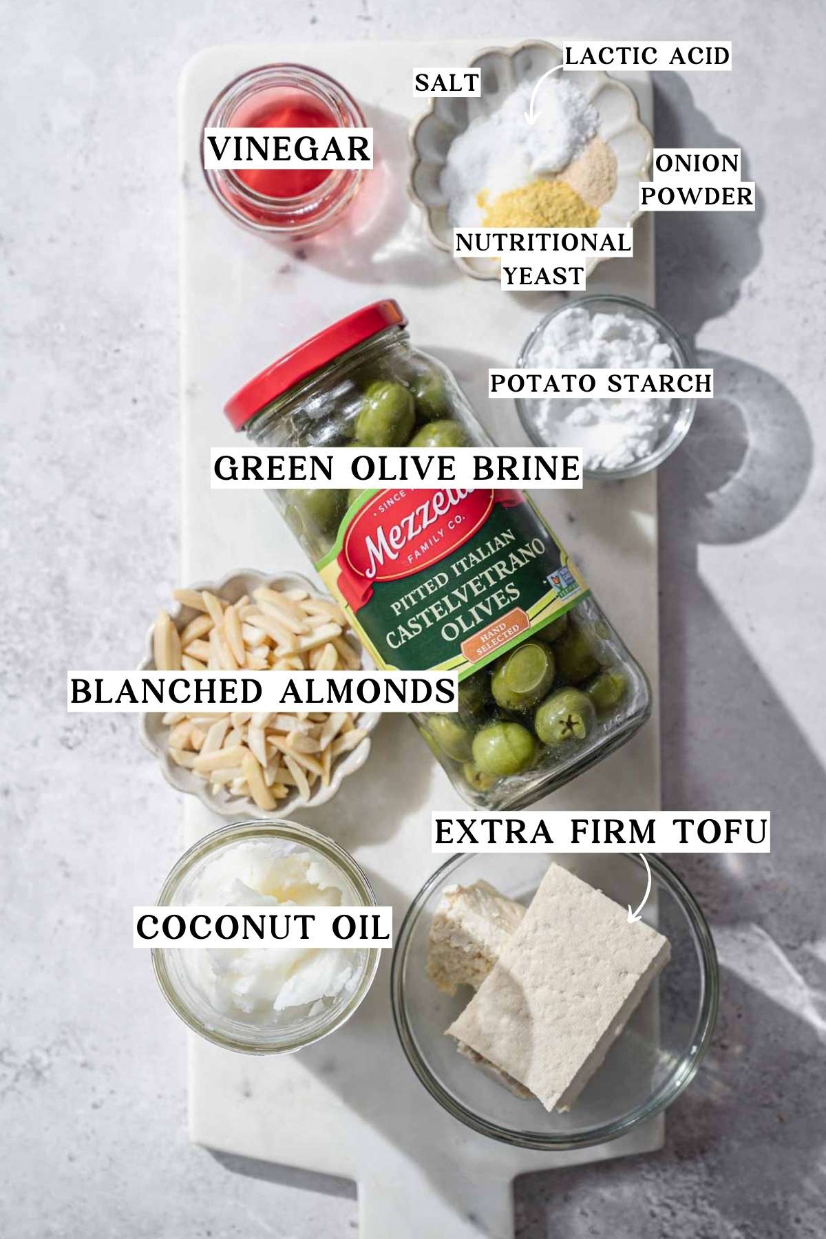Ingredients arranged on a marble surface, including tofu, almonds, coconut oil, olive brine, potato starch, vinegar, and seasonings.