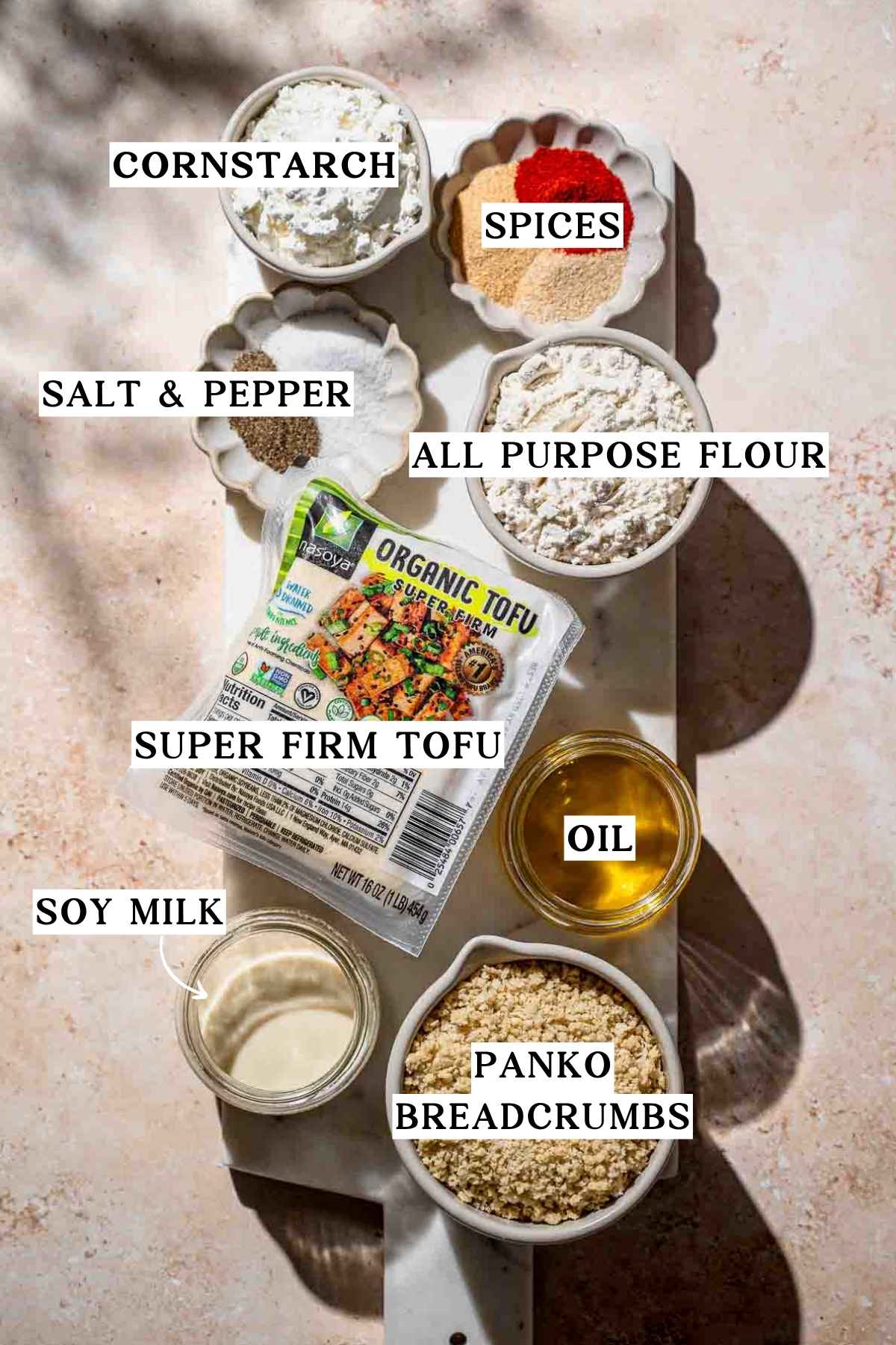 ingredients like super firm tofu, panko breadcrumbs, oil, spices and flour on a marble cutting board.
