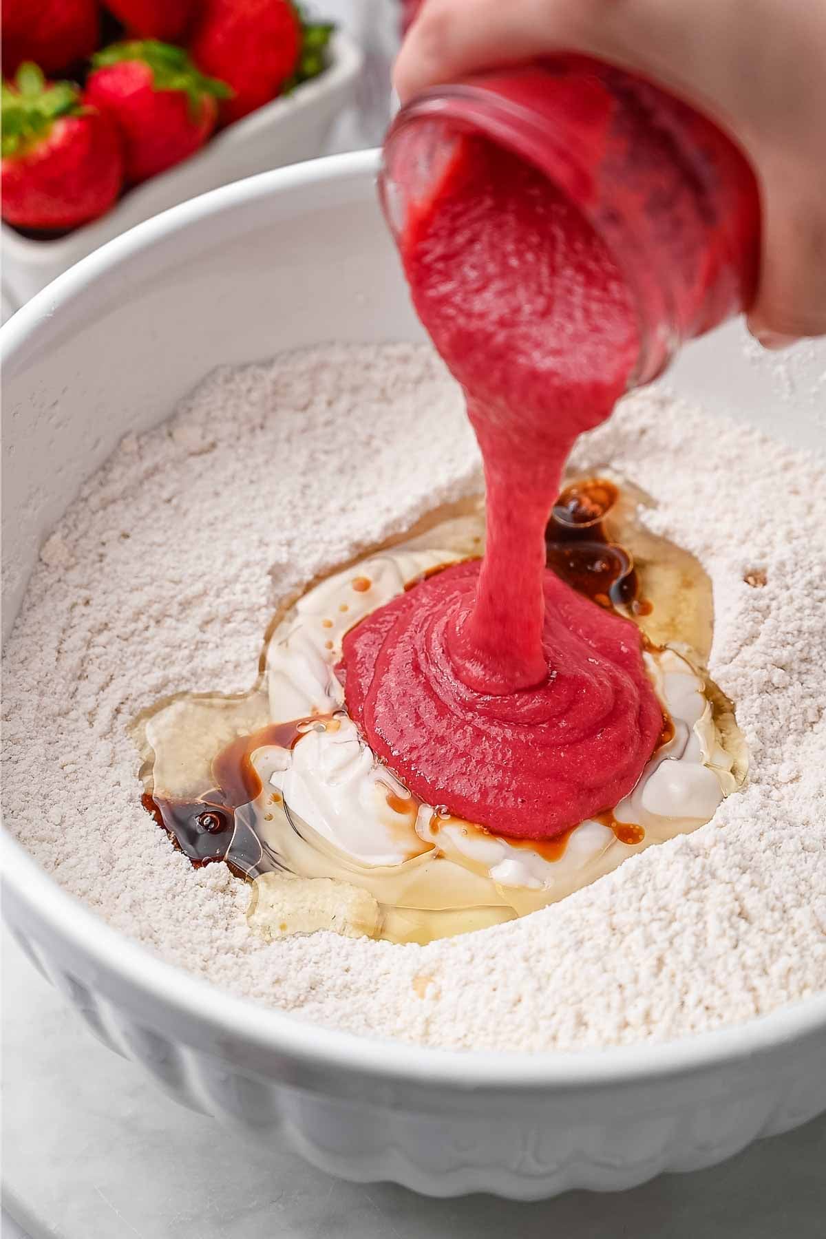 a hand pouring the strawberry puree on the wet ingredients into the mixing bowl