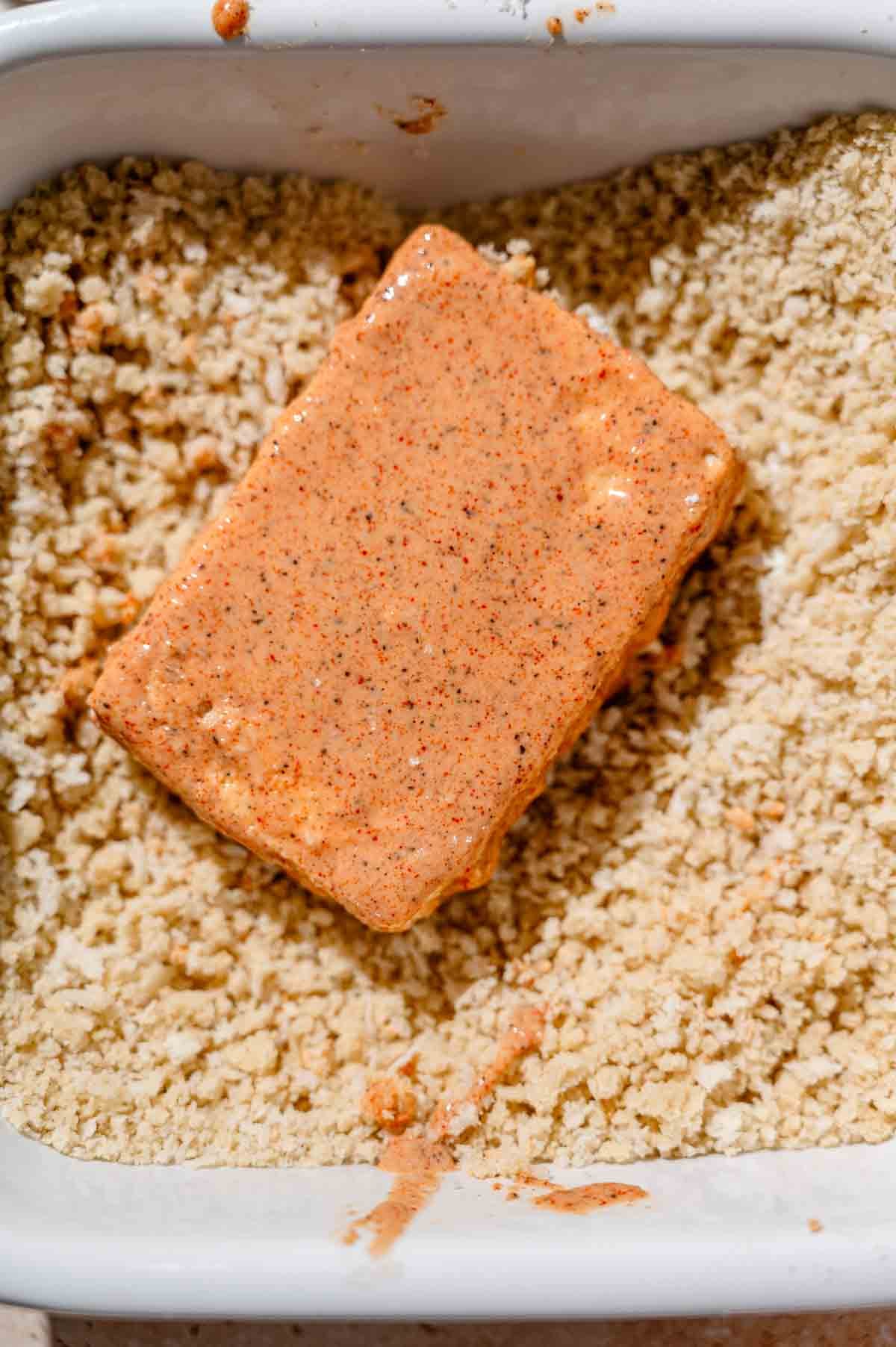 a batter coated piece of tofu in a dish filled with panko breadcrumbs.