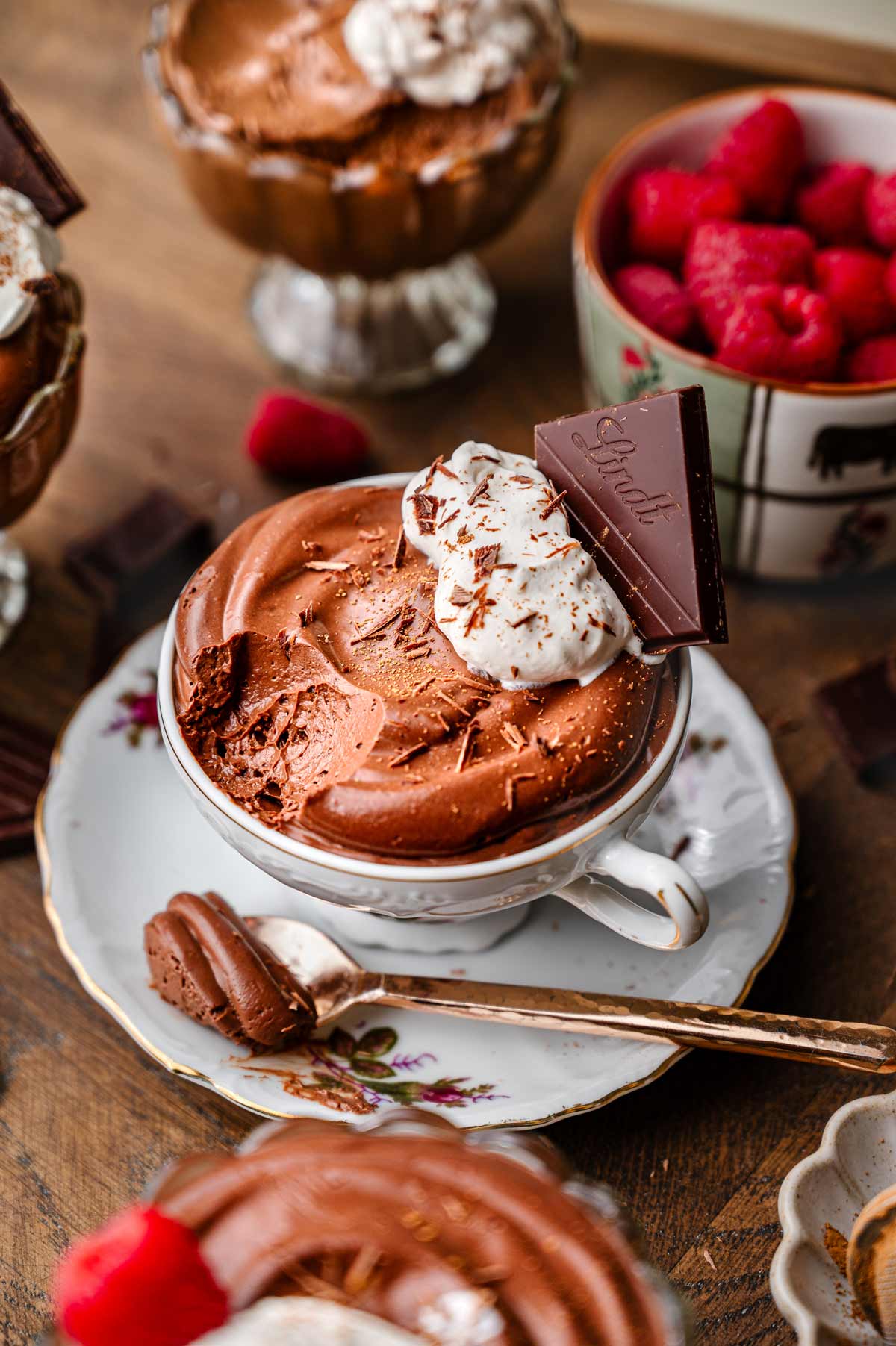 Chocolate vegan mousse served in a teacup with dairy-free whipped cream, shaved chocolate, raspberries, and dark chocolate.