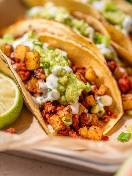 Close-up of vegan chorizo tacos with crispy potatoes, guacamole, cilantro crema, and scallions in corn tortillas.