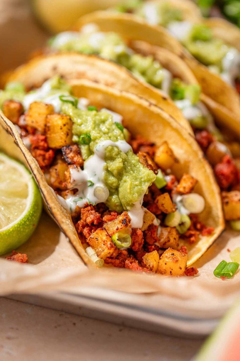 Close-up of vegan chorizo tacos with crispy potatoes, guacamole, cilantro crema, and scallions in corn tortillas.