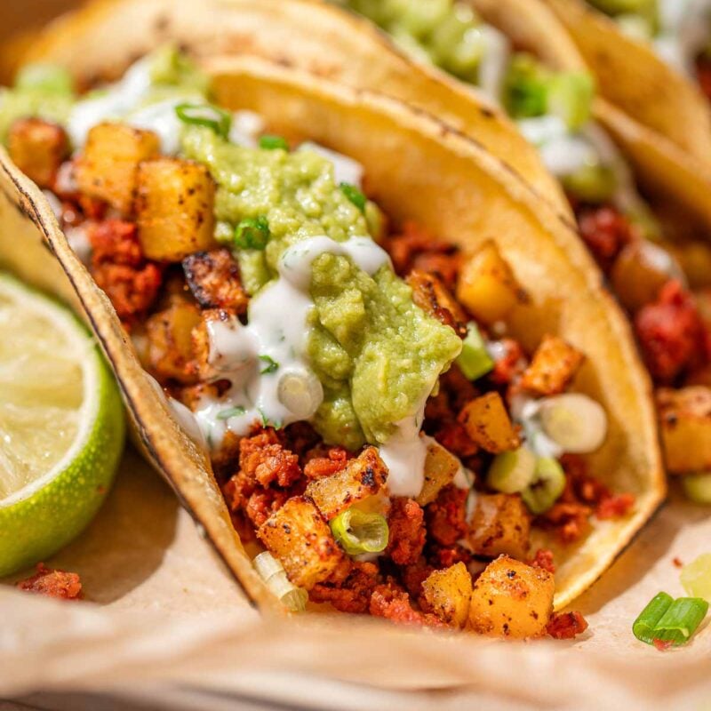 Close-up of vegan chorizo tacos with crispy potatoes, guacamole, cilantro crema, and scallions in corn tortillas.