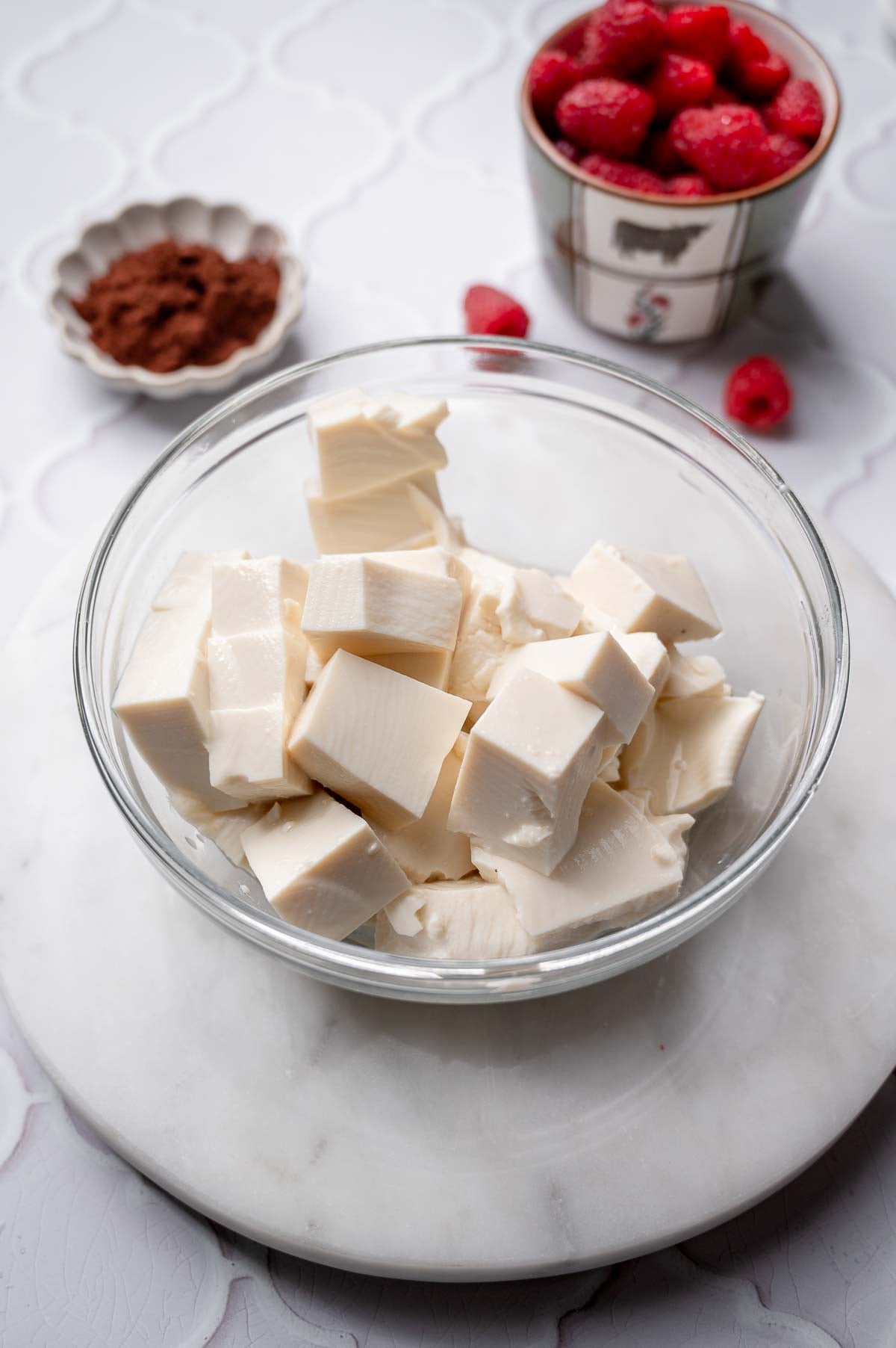 Cubed silken tofu in a glass bowl with cocoa powder and raspberries in the background.
