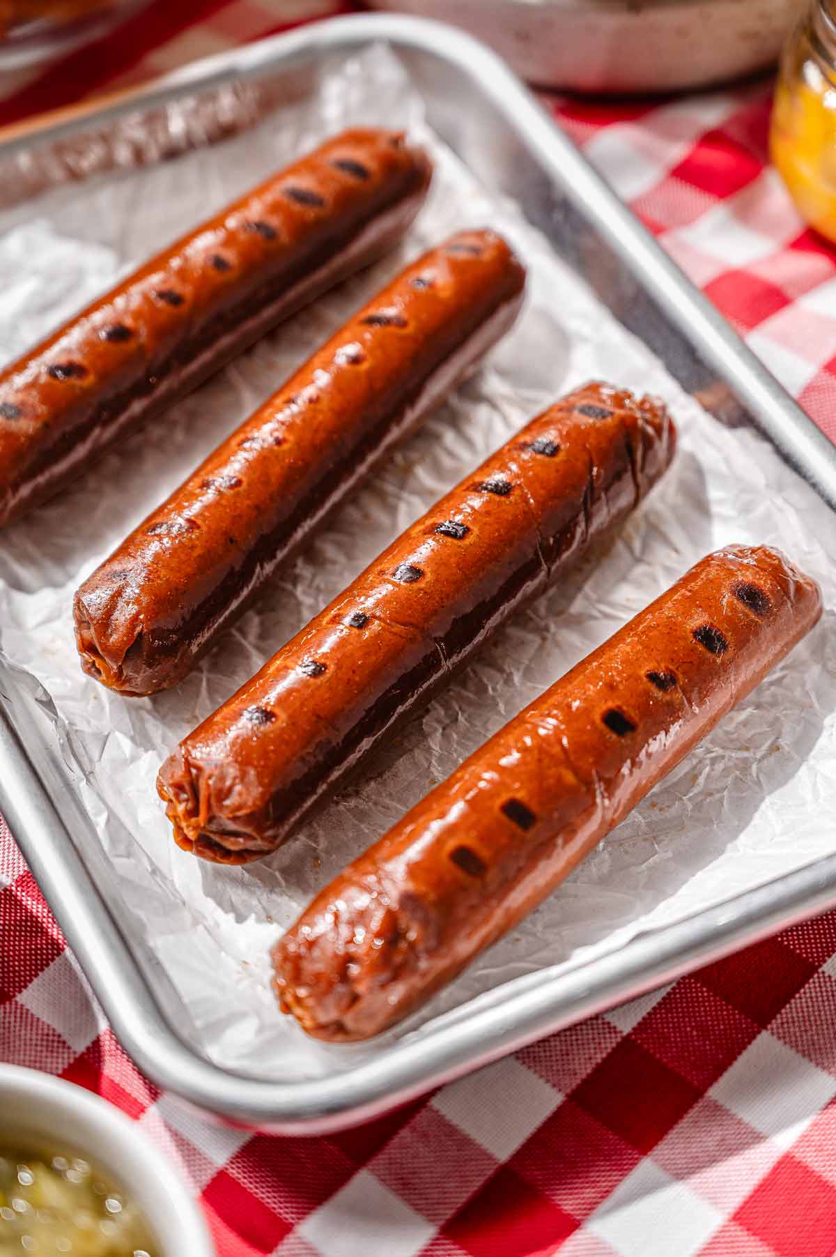 Close-up of four homemade vegan hot dogs with grill marks on a parchment-lined tray set on a red gingham tablecloth.