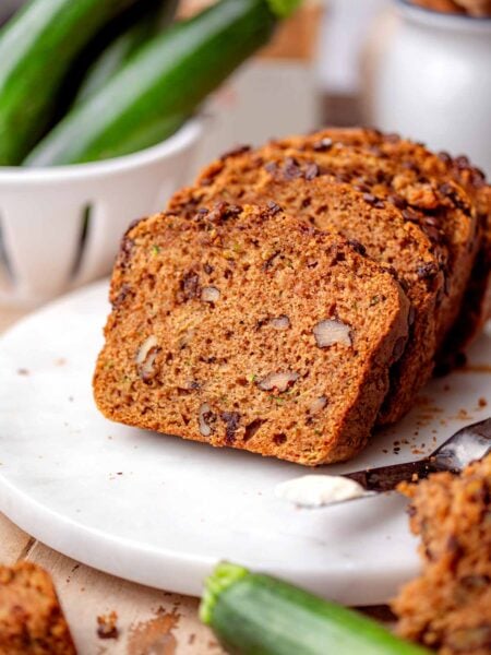 Vegan chocolate chip zucchini bread sliced on a white plate with walnuts and fresh zucchini in the background.