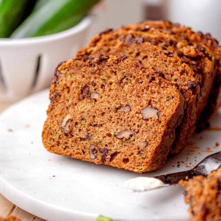 Vegan chocolate chip zucchini bread sliced on a white plate with walnuts and fresh zucchini in the background.