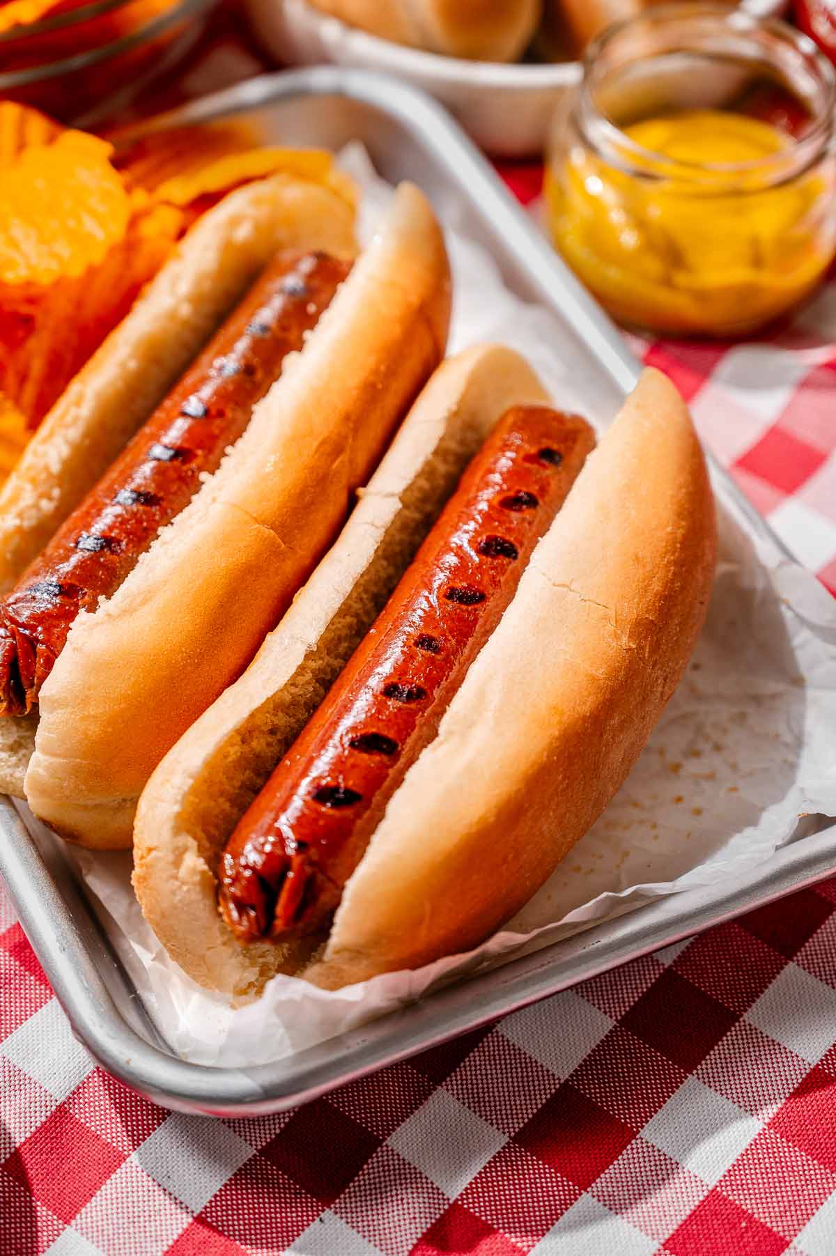 Vegan hot dogs nestled in soft buns on a tray with chips, mustard, and relish, styled on a red checkered picnic tablecloth.