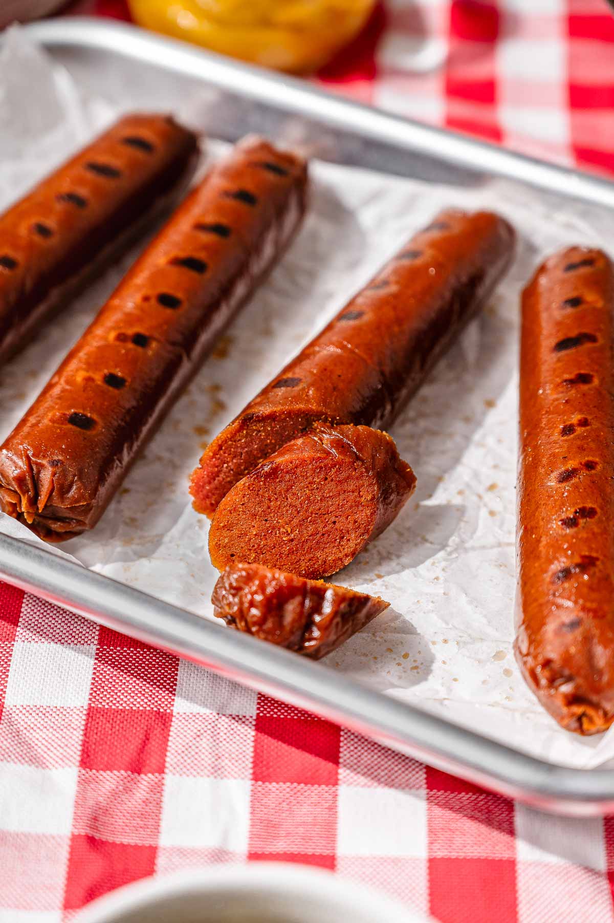 Sliced vegan hot dog showing the firm seitan interior texture, surrounded by whole grilled hot dogs on a parchment-lined tray.
