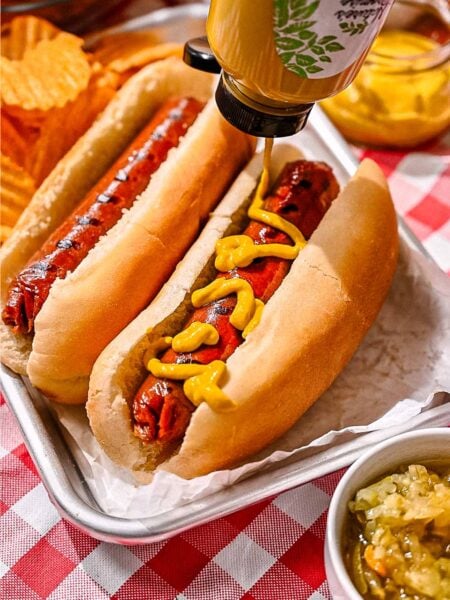 Mustard being squeezed onto a grilled vegan hot dog in a bun, served on a metal tray with relish and potato chips.