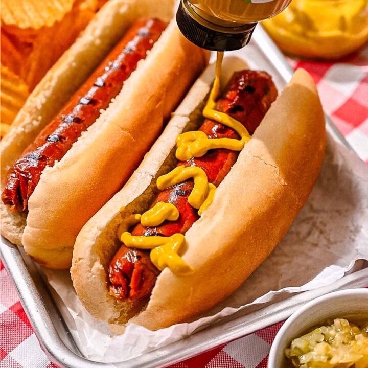 Mustard being squeezed onto a grilled vegan hot dog in a bun, served on a metal tray with relish and potato chips.