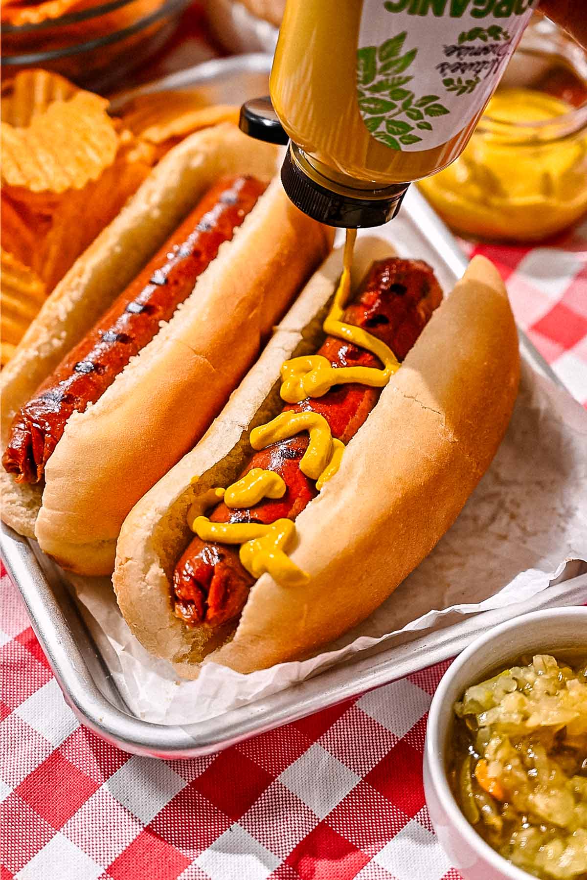 Mustard being squeezed onto a grilled vegan hot dog in a bun, served on a metal tray with relish and potato chips.
