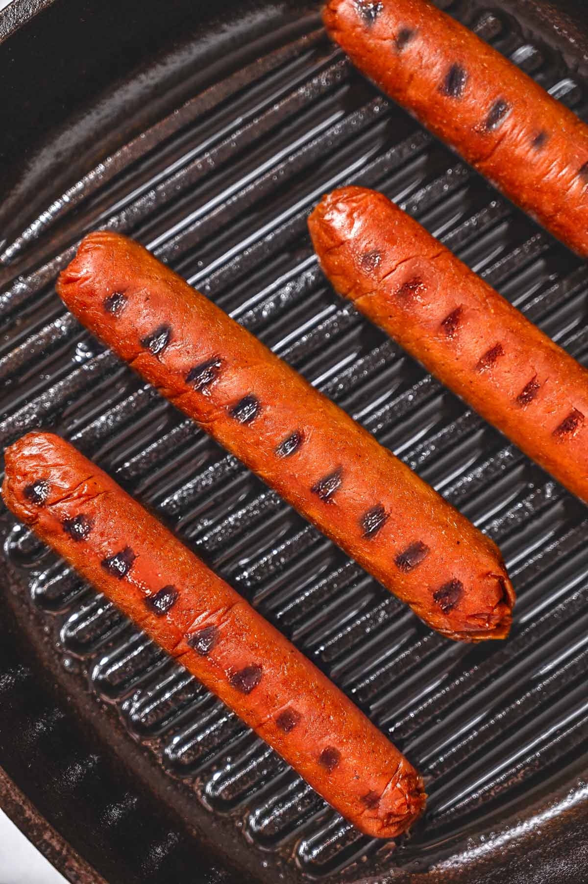 Three homemade vegan hot dogs with char marks cooking on a cast iron grill pan.