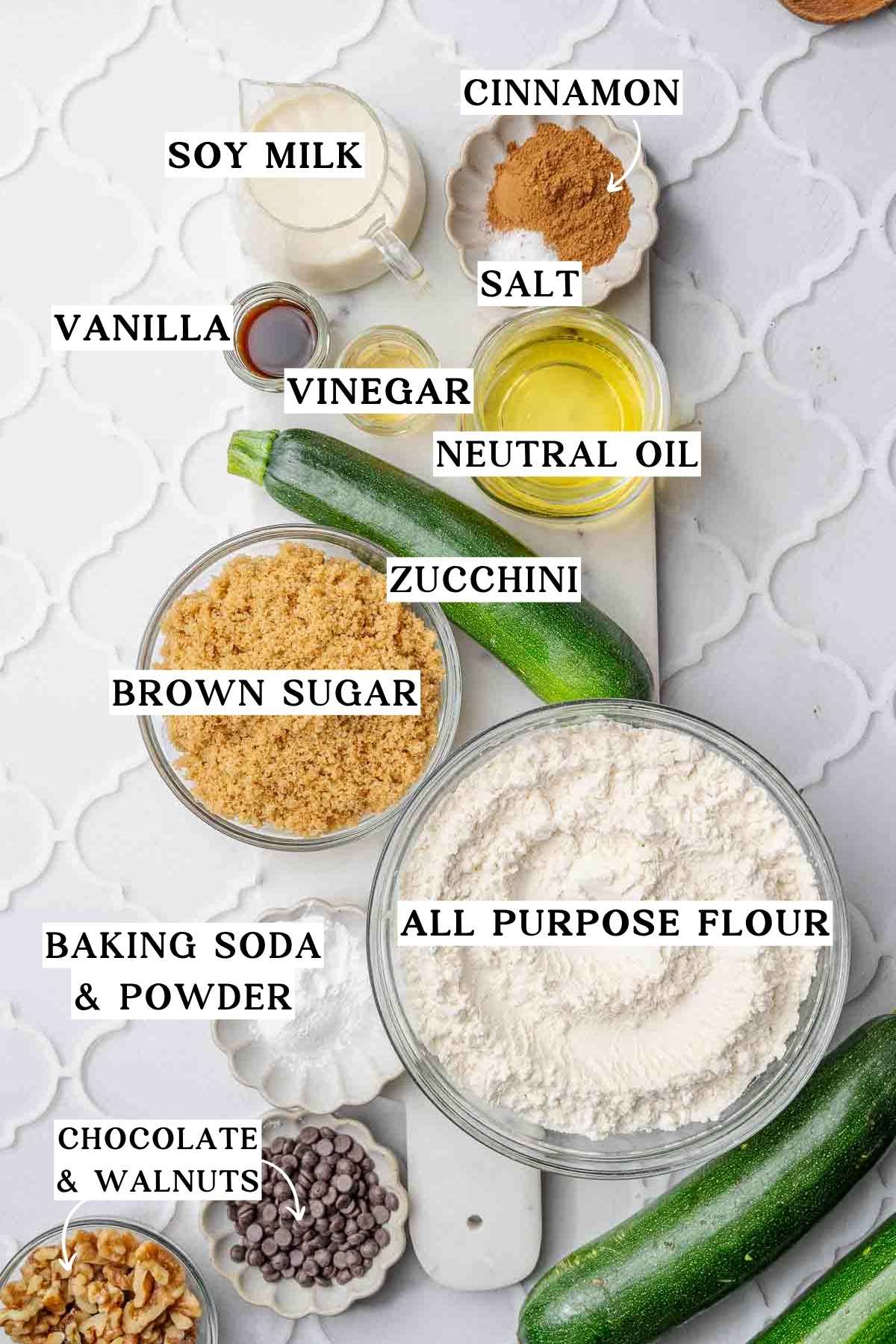 Ingredients for vegan zucchini bread including zucchini, flour, brown sugar, soy milk, oil, cinnamon, vanilla, vinegar, walnuts and chocolate chips.