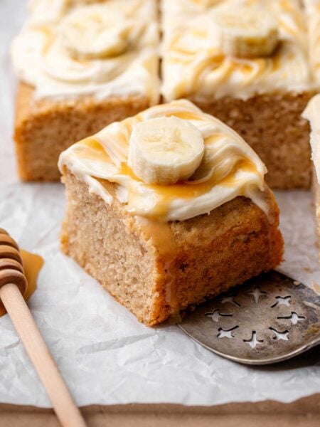 Thick square of moist vegan banana cake topped with cream cheese frosting, caramel drizzle, and a fresh banana slice.