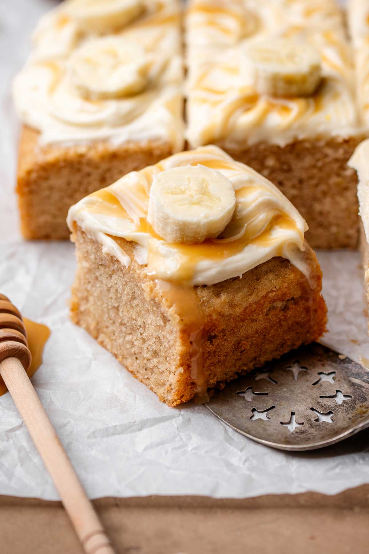 Thick square of moist vegan banana cake topped with cream cheese frosting, caramel drizzle, and a fresh banana slice.