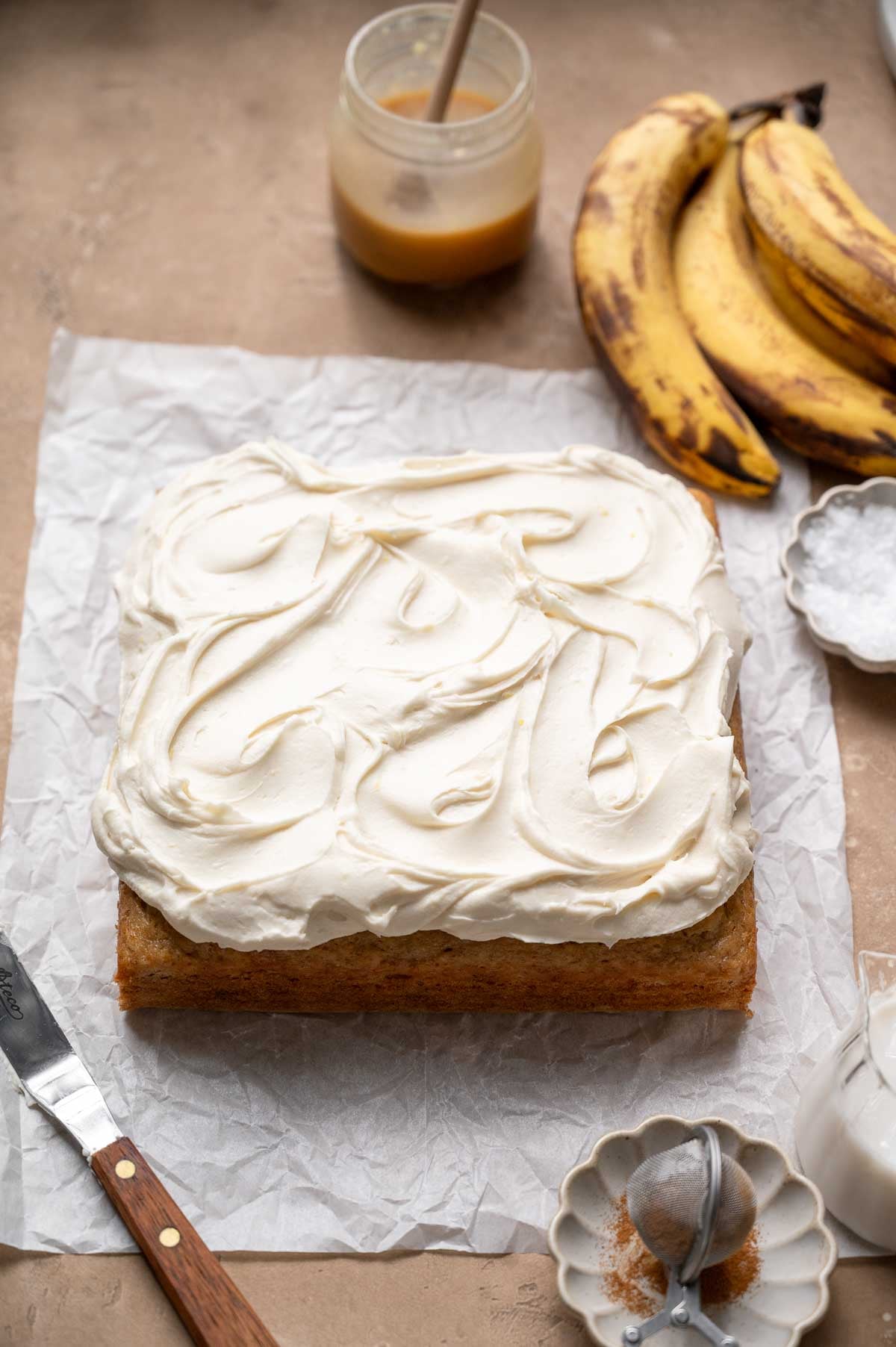 Square vegan banana cake covered with a thick layer of swirled cream cheese frosting.