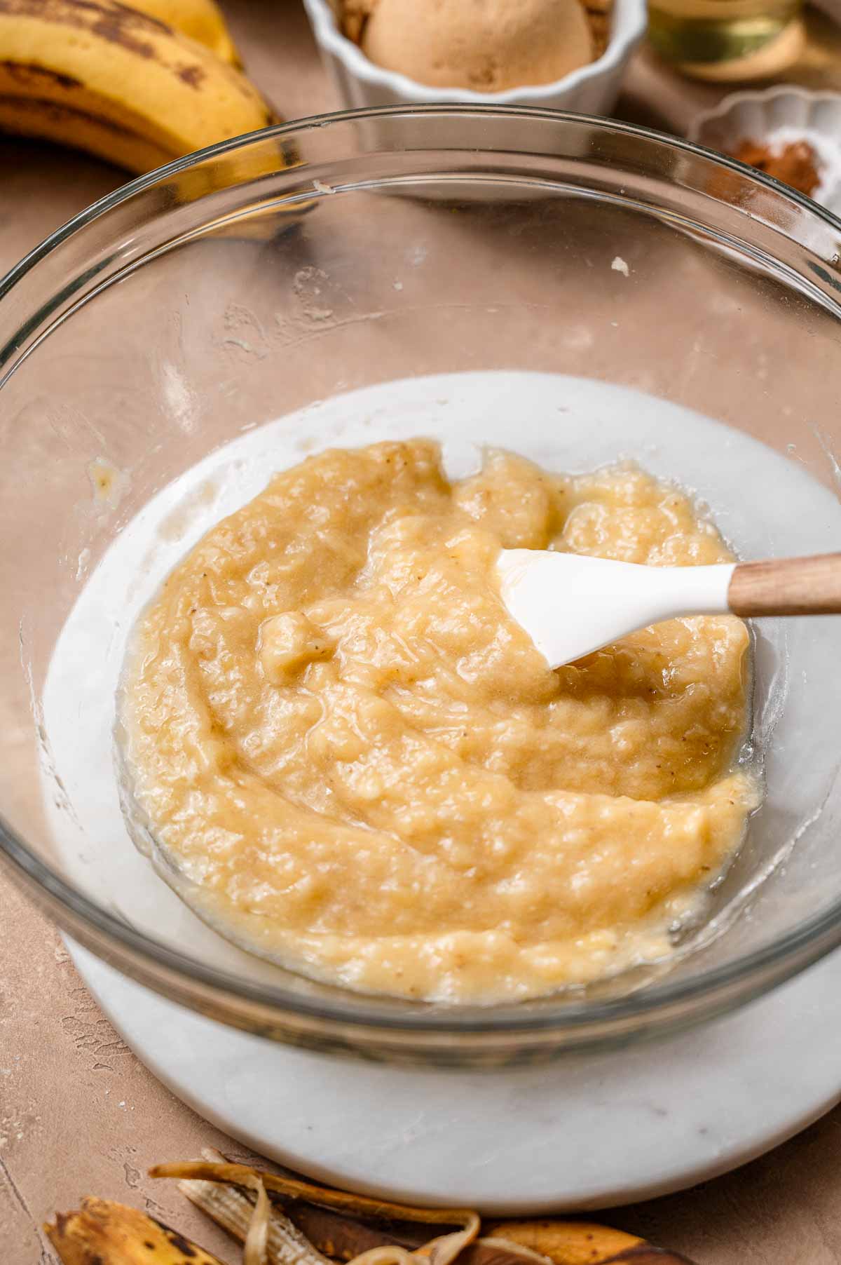 Mashed ripe bananas in a glass mixing bowl with a white spatula.