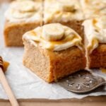 Moist vegan banana cake square topped with cream cheese frosting, caramel drizzle, and a fresh banana slice.
