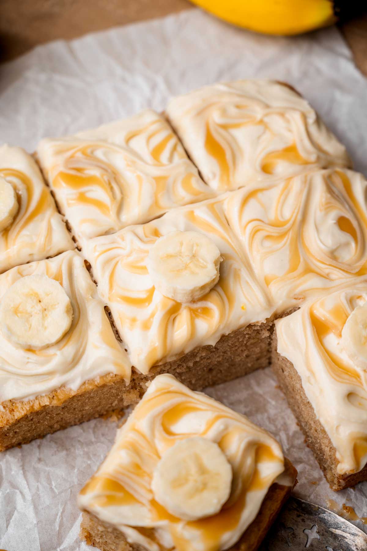 Sliced vegan banana cake topped with swirled cream cheese frosting, caramel, and fresh banana slices.