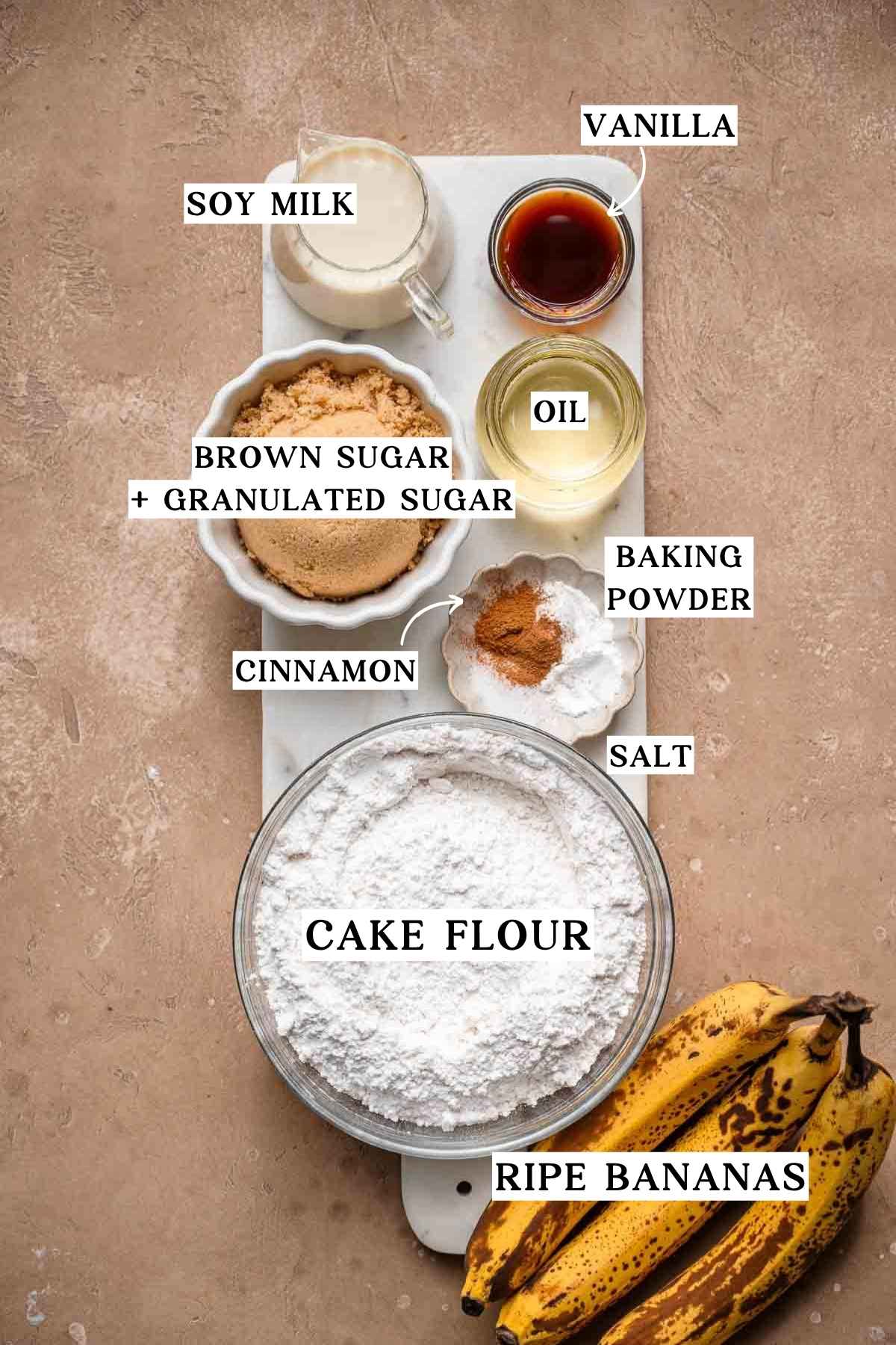 Overhead view of labeled vegan banana cake ingredients, including ripe bananas, cake flour, sugars, soy milk, oil, vanilla, cinnamon, salt, and baking powder.