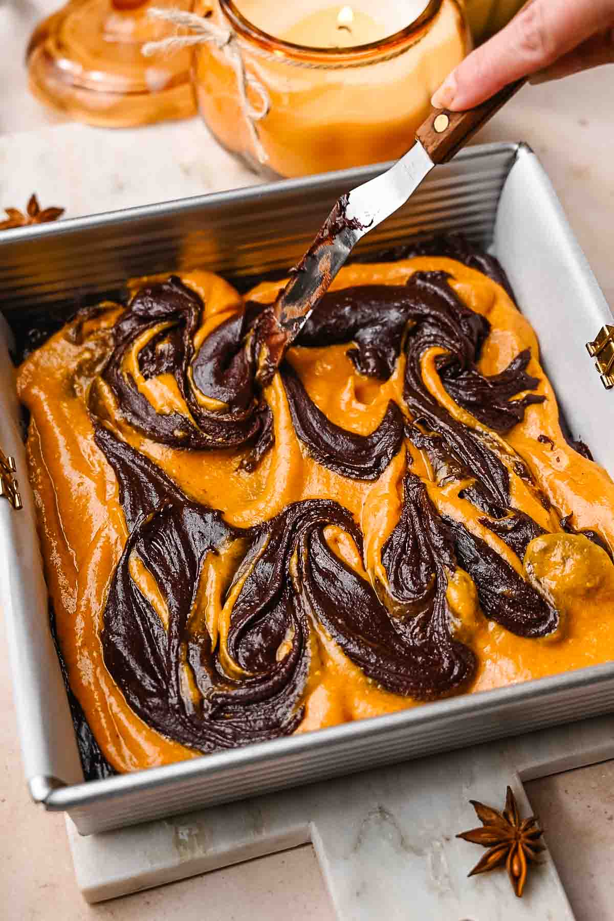 a hand using a knife swirling the pumpkin cheesecake filling into the vegan brownie batter.