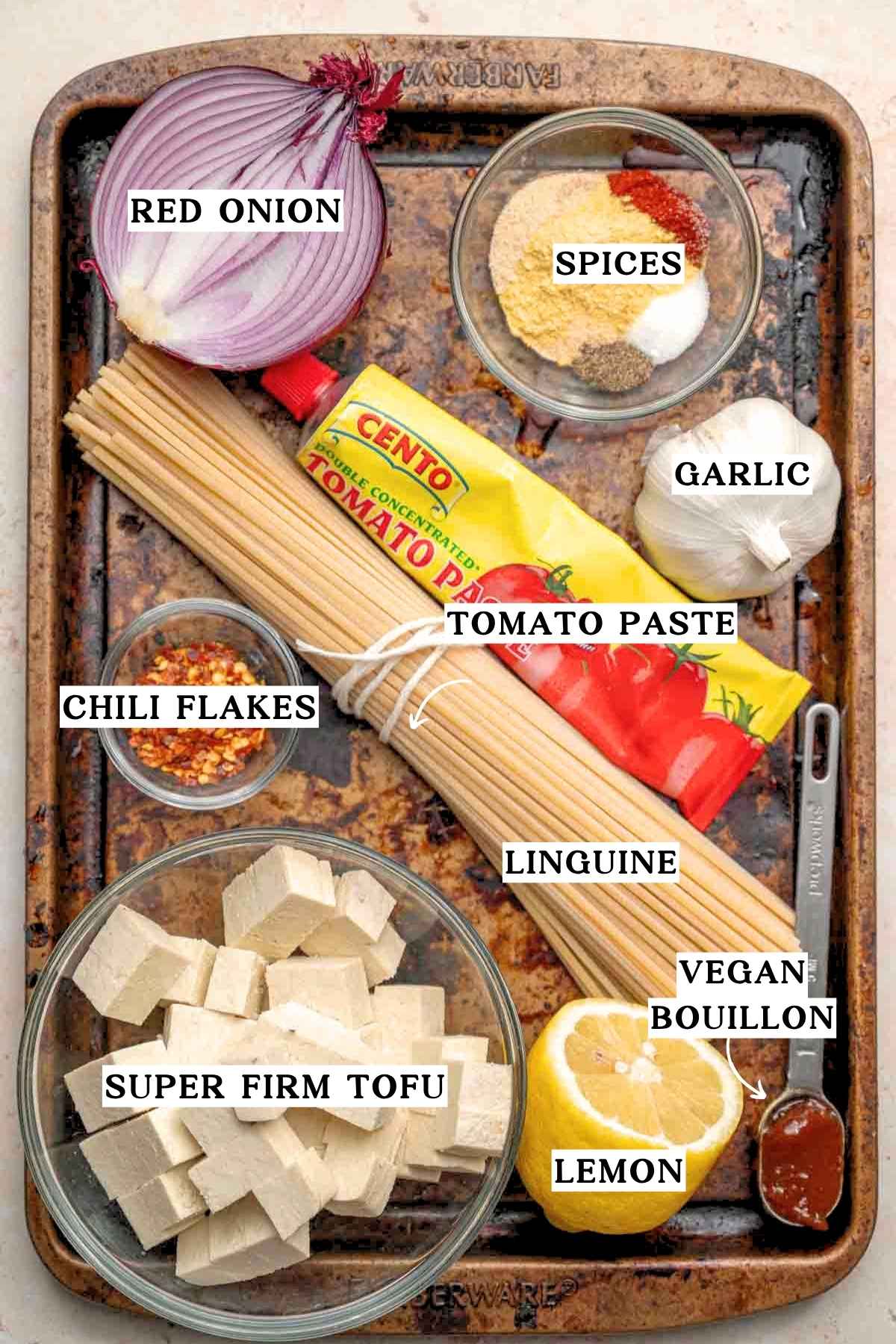 ingredients like dry linguine, tomato paste, red onion, garlic and chili flakes on a metal baking tray.
