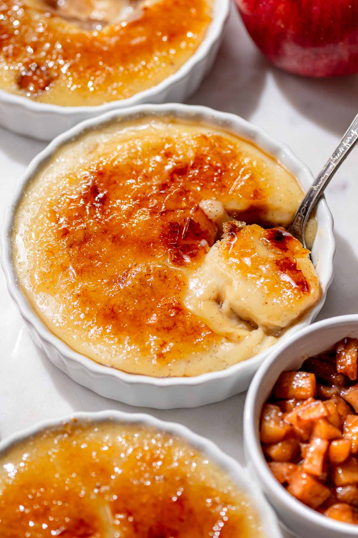 Apple pie crème brûlée with a golden caramelized sugar crust and a spoonful of creamy custard, surrounded by apples.