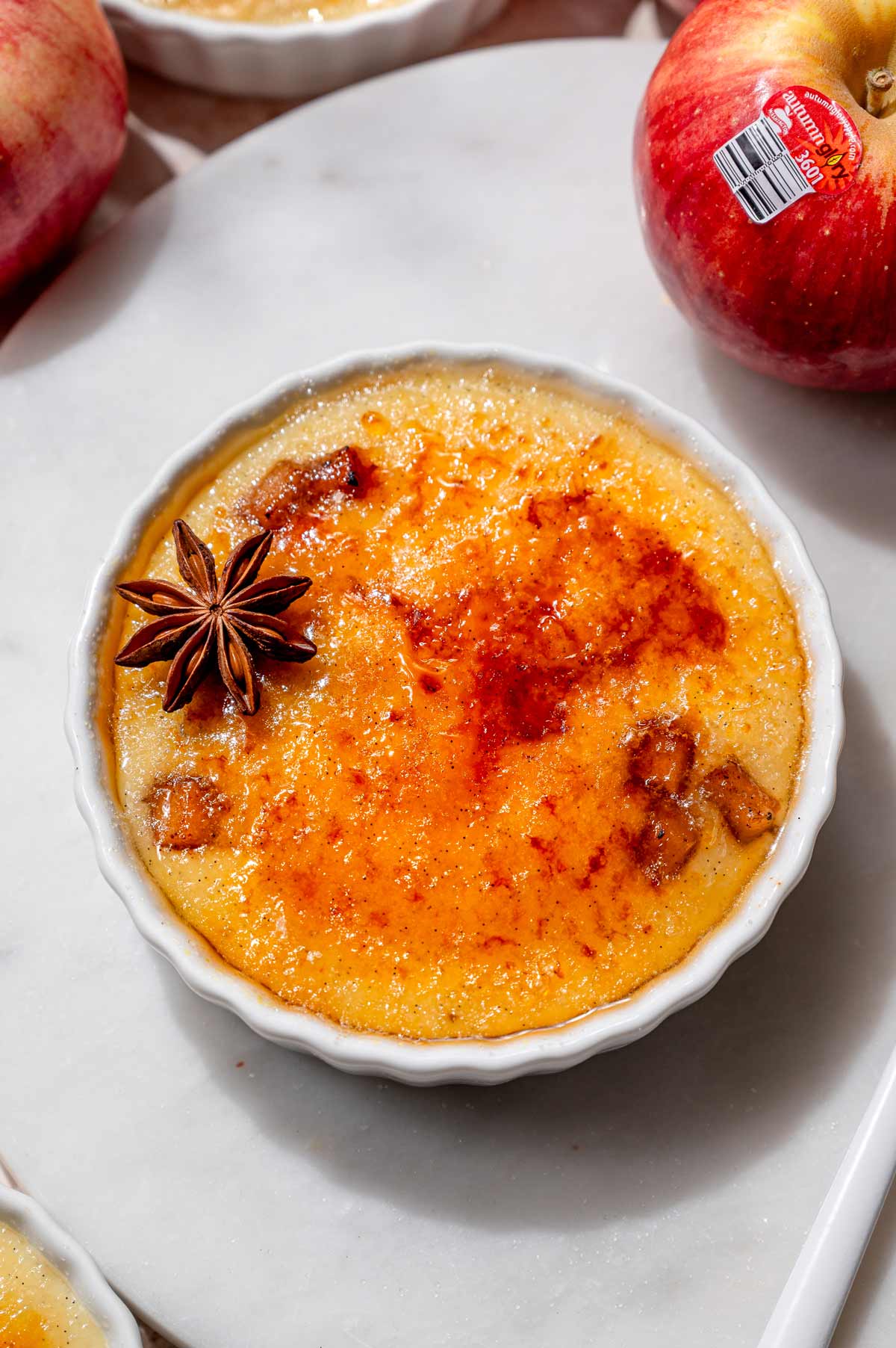 Finished apple crème brûlée with a crisp, golden sugar topping, garnished with star anise and surrounded by fresh red apples.