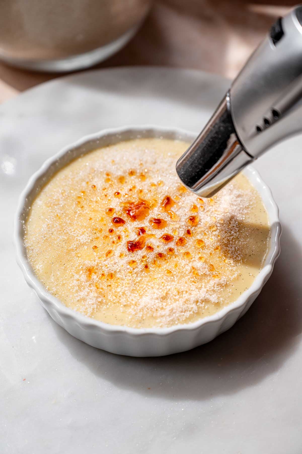 A kitchen torch melting and browning a thin layer of sugar over custard in a white ramekin.