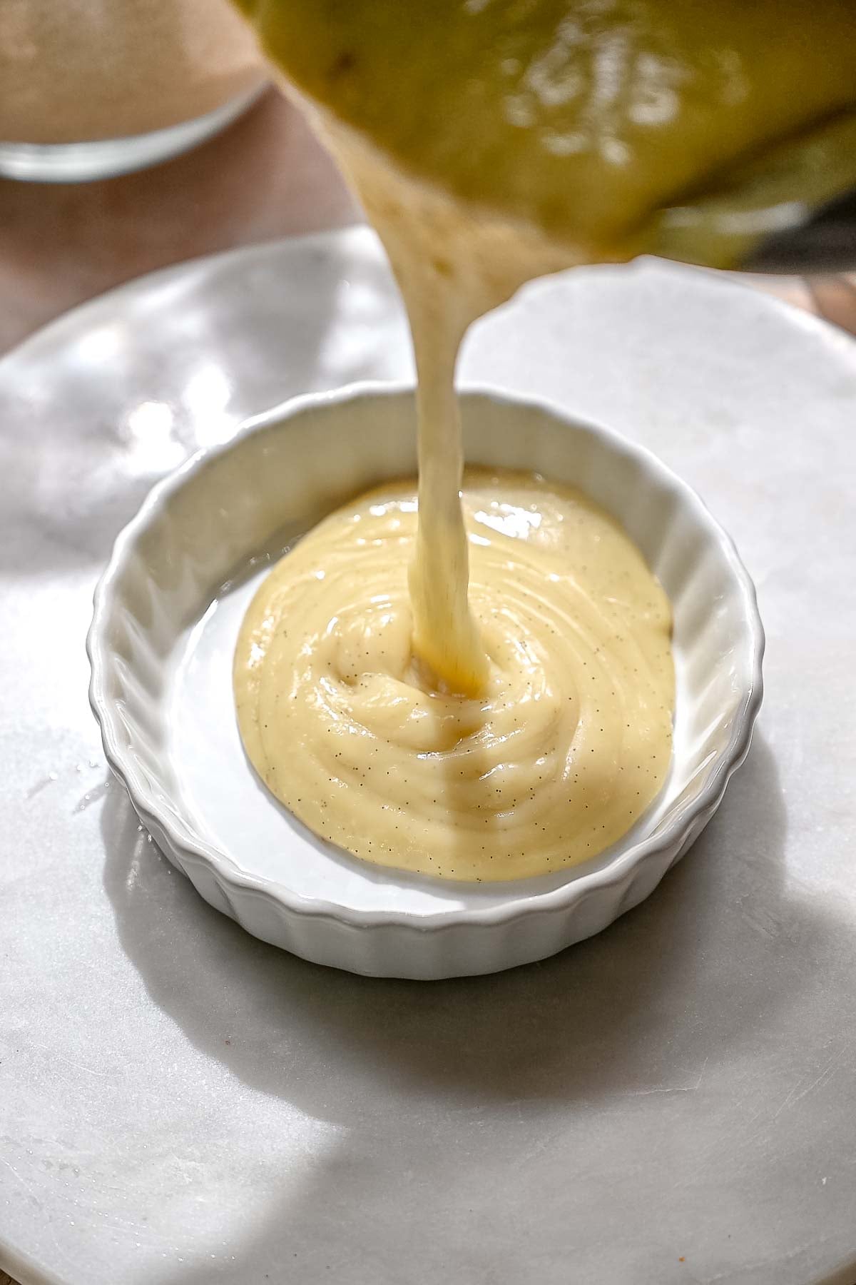 Warm, creamy custard being poured into a small white fluted ramekin.