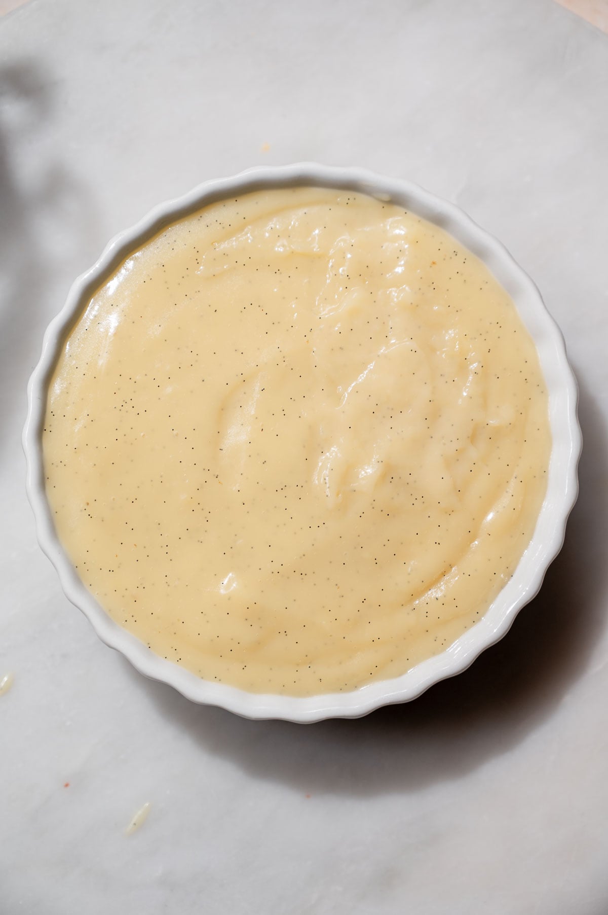 Smooth, pale yellow vanilla bean custard dotted with vanilla seeds in a white ceramic ramekin.