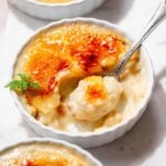 White ramekins filled with vegan crème brûlée, with a spoon scooping through the crisp caramelized sugar crust of one.