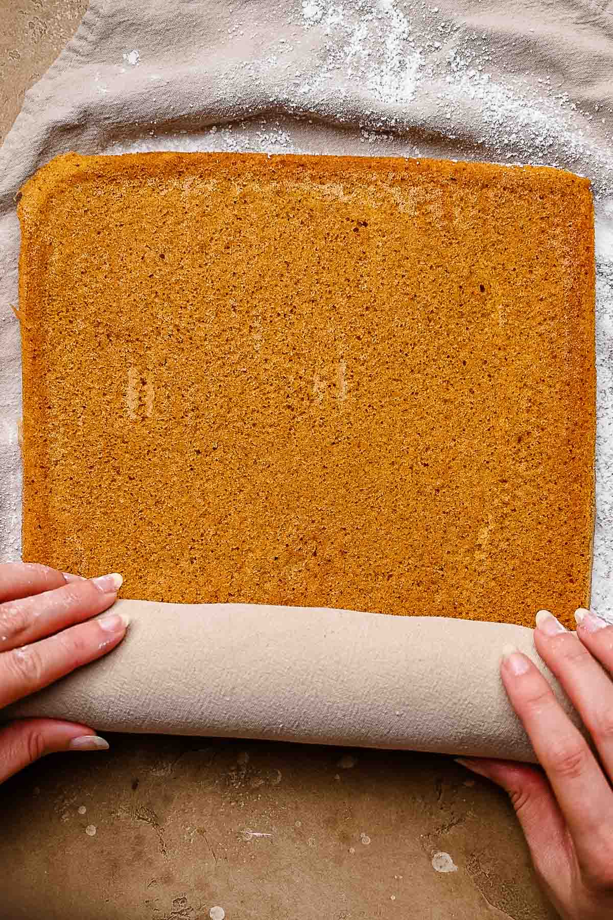 Hands rolling a rectangular pumpkin sponge cake in a kitchen towel to help it hold its shape.