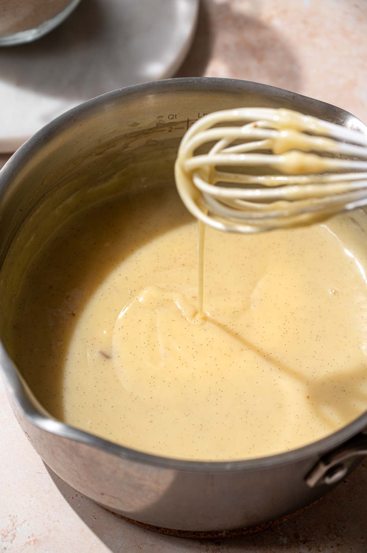 Thickened eggless vanilla custard dripping from a whisk into a stainless steel saucepan.