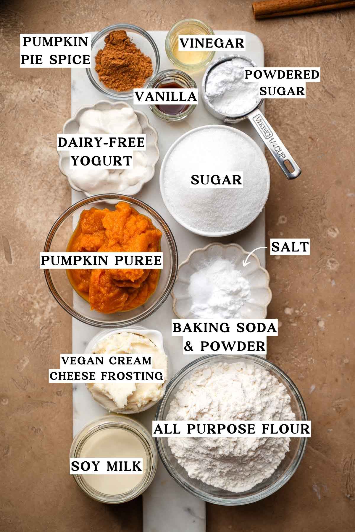 Overhead view of pumpkin roll ingredients including pumpkin puree, soy milk, dairy-free yogurt, flour, spices, and frosting.